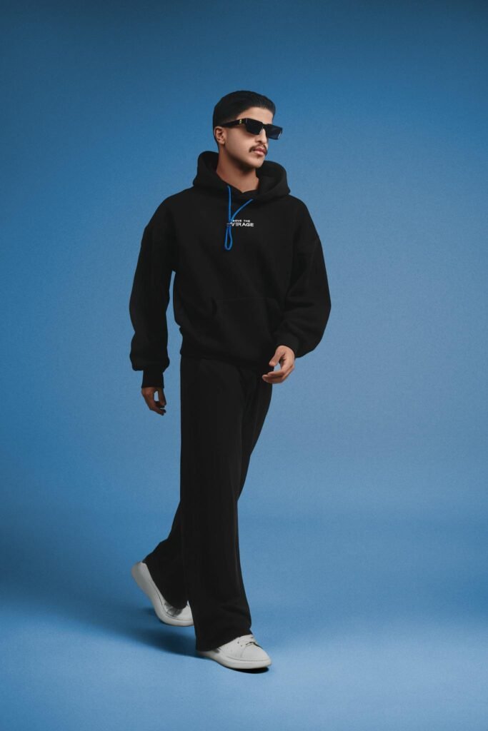 Fashionable man in black hoodie and pants walking against a blue backdrop.