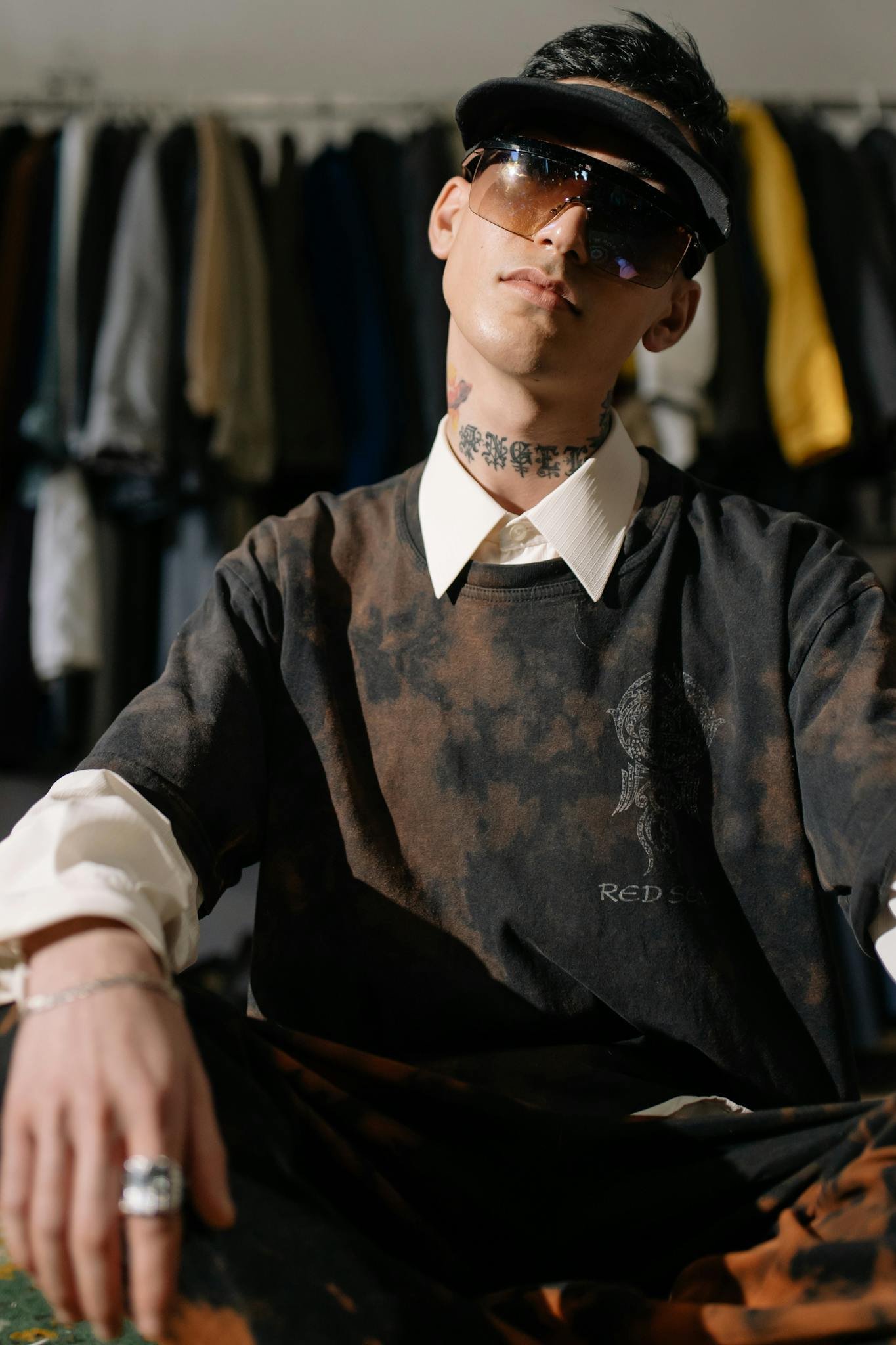 Stylish man posing with tattoos and sunglasses in front of a clothes rack indoors.